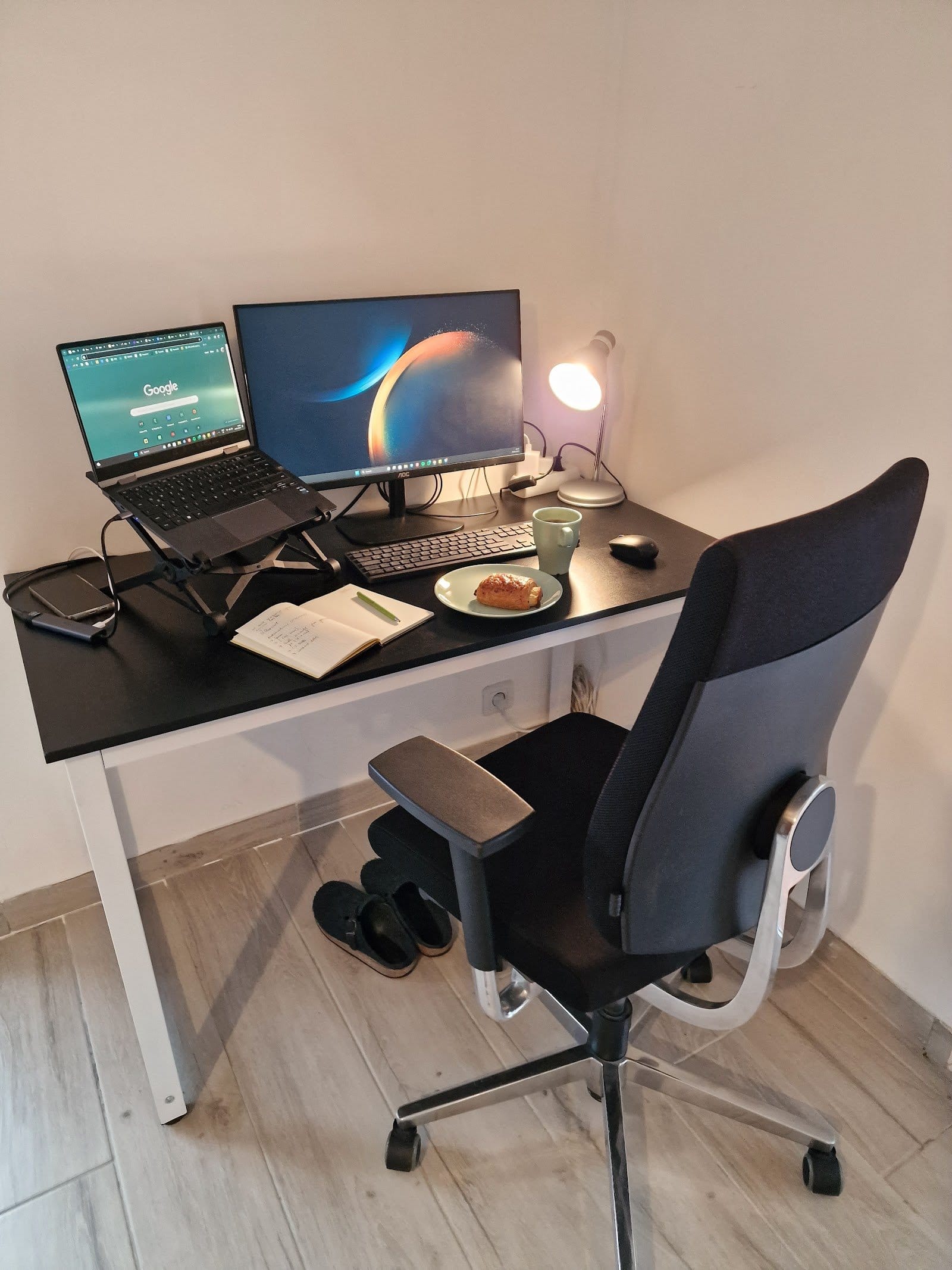 Small workspace with desk, laptop on stand, monitor, lamp and chair in bright room.