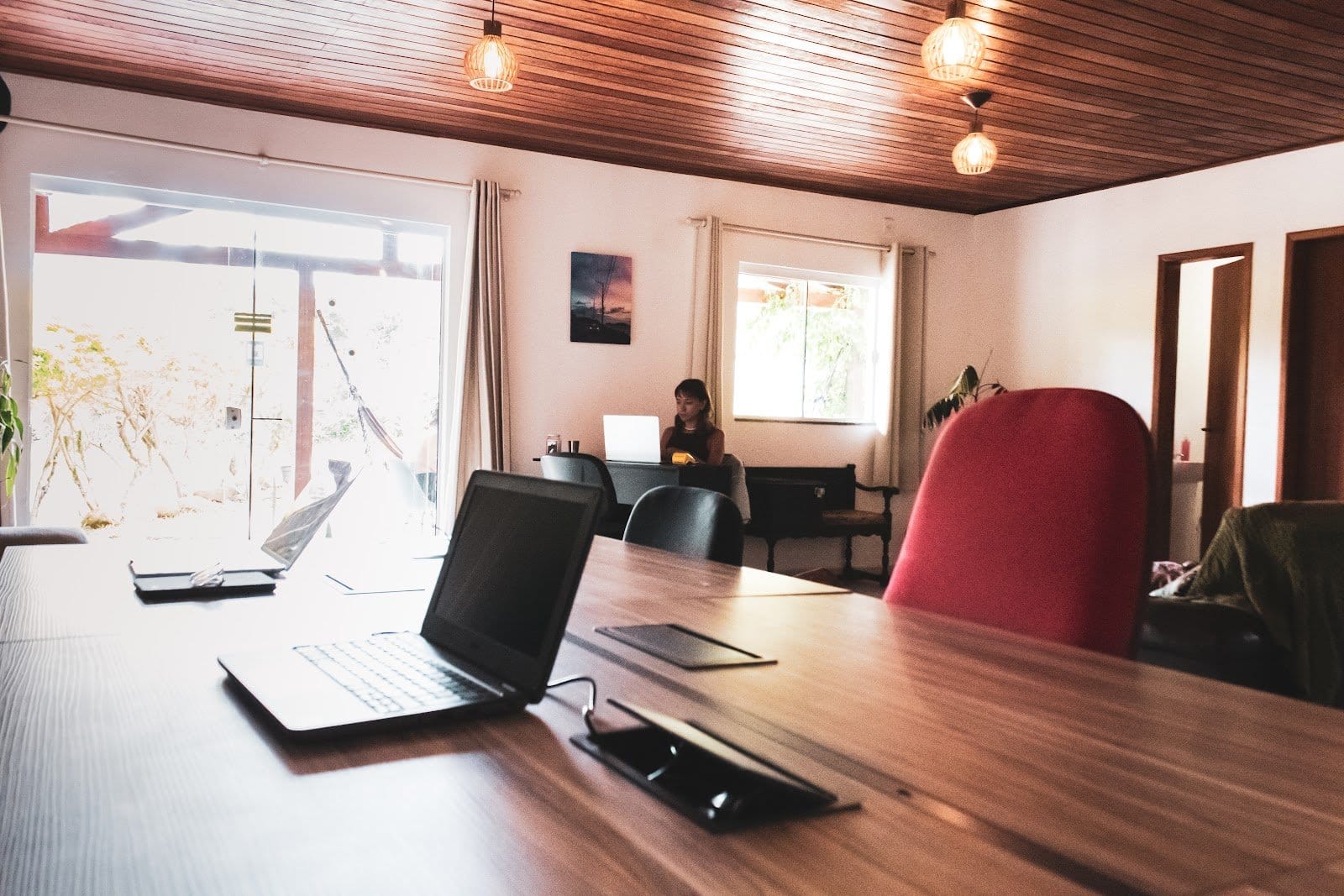 Bright coworking room with laptops on a long desk and a guest working by the window at Nômades Riverside, Florianópolis