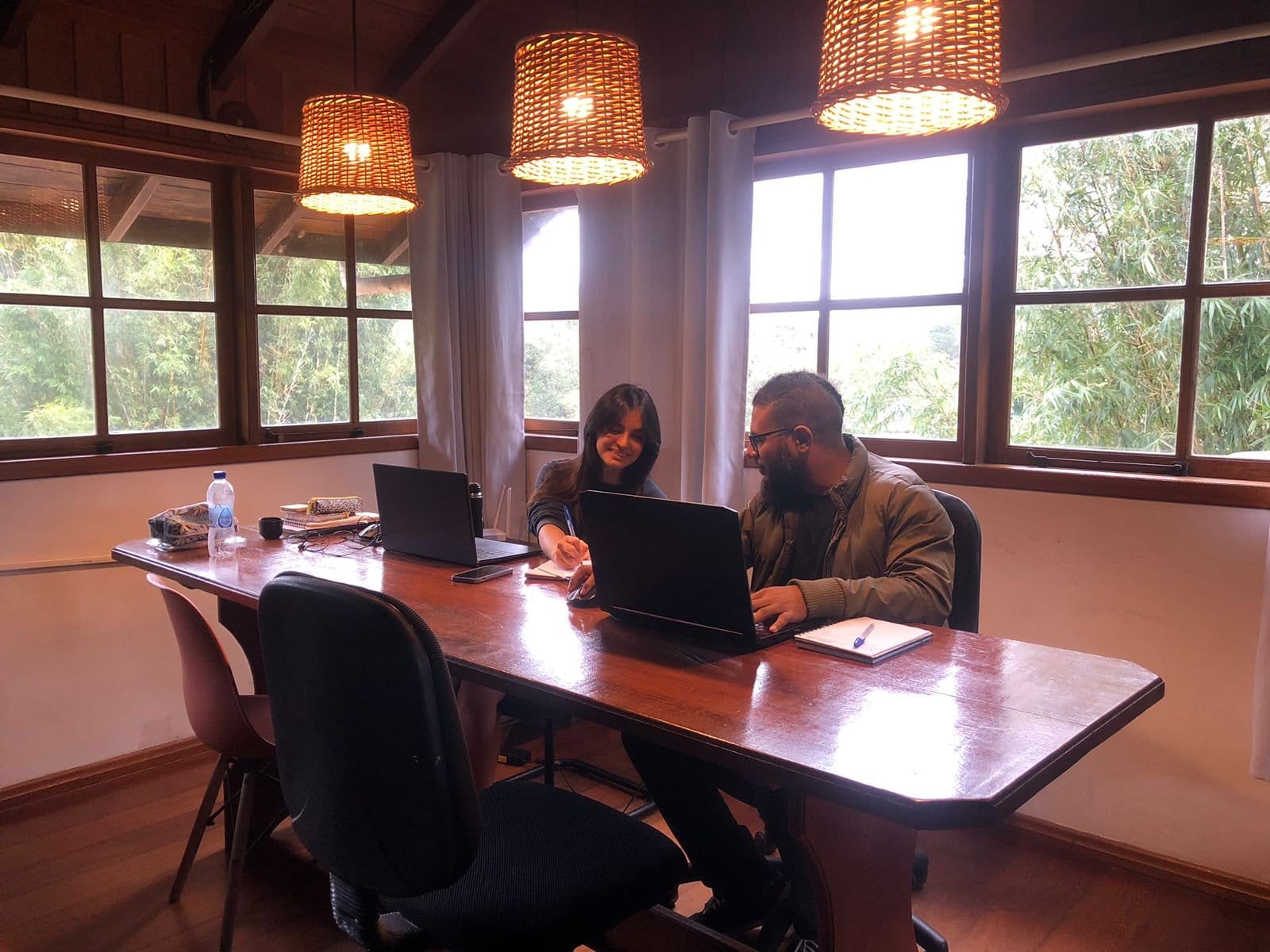 Two guests working on laptops at a wooden table under woven pendant lights in the coworking room at Nômades Riverside, Florianópolis