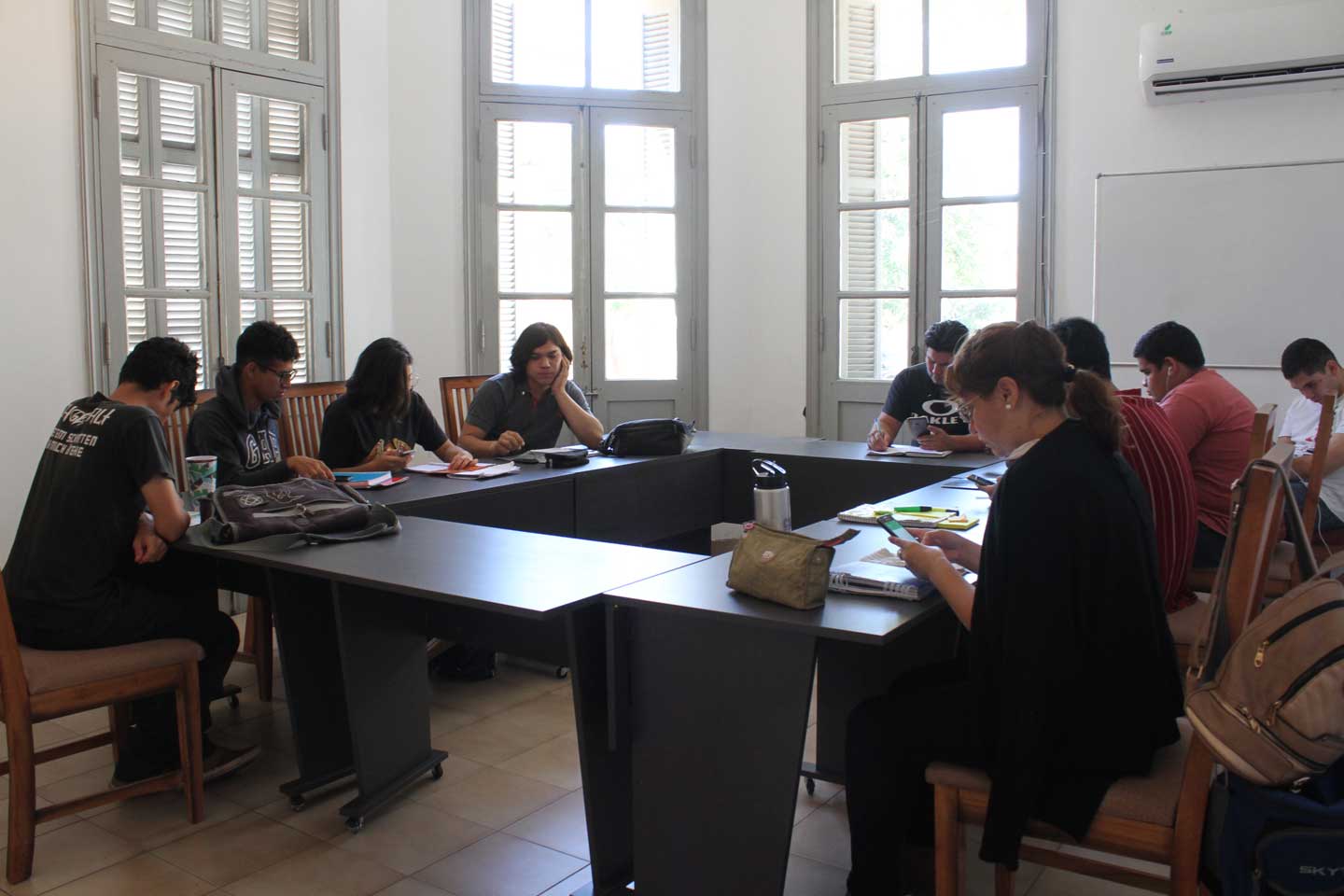 Room with large windows and shutters, several people seated at desks working or using phones, bright natural light.