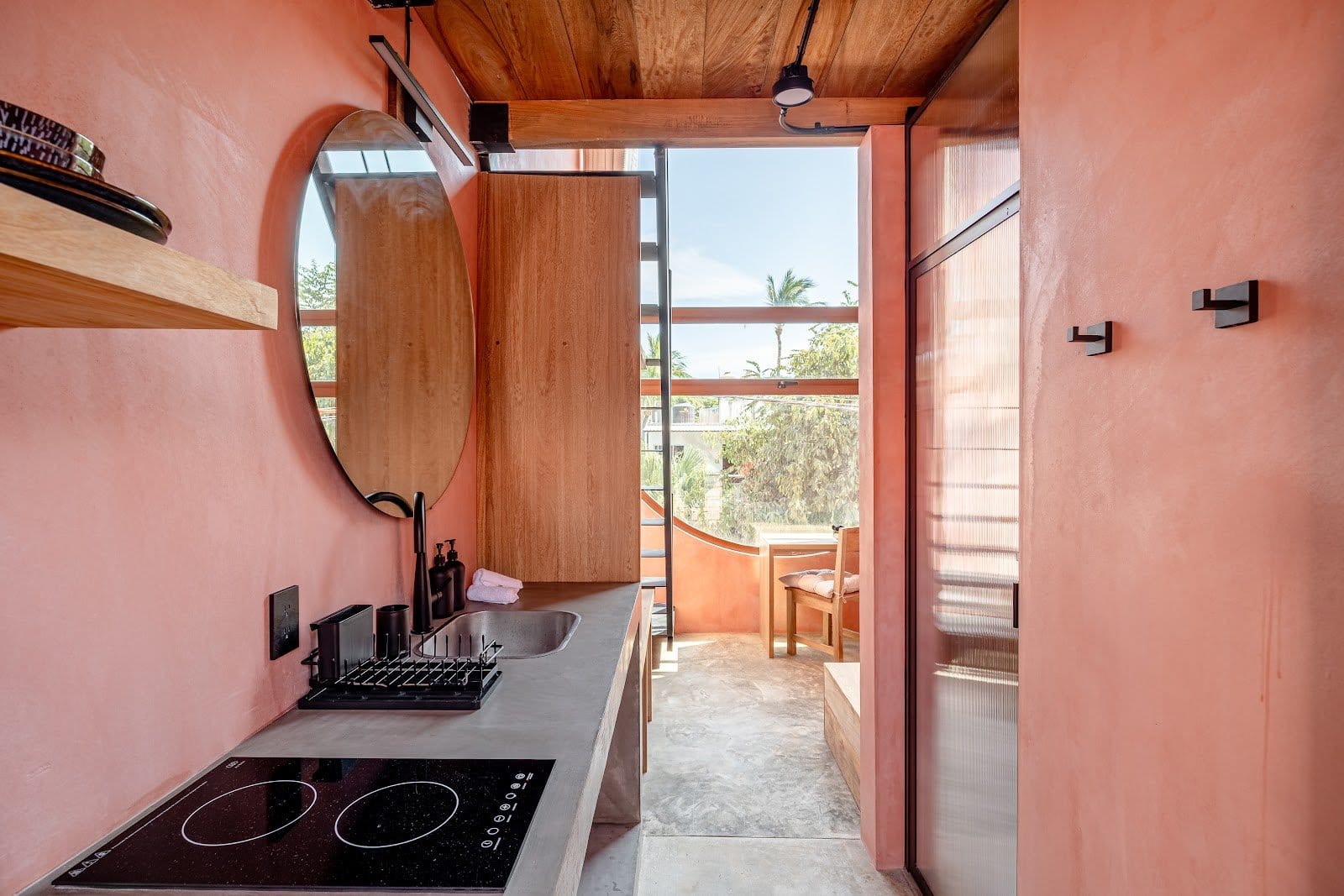 In-room kitchenette at Amplitude Coliving with a cooktop, round mirror, wood cabinets, and balcony doors.