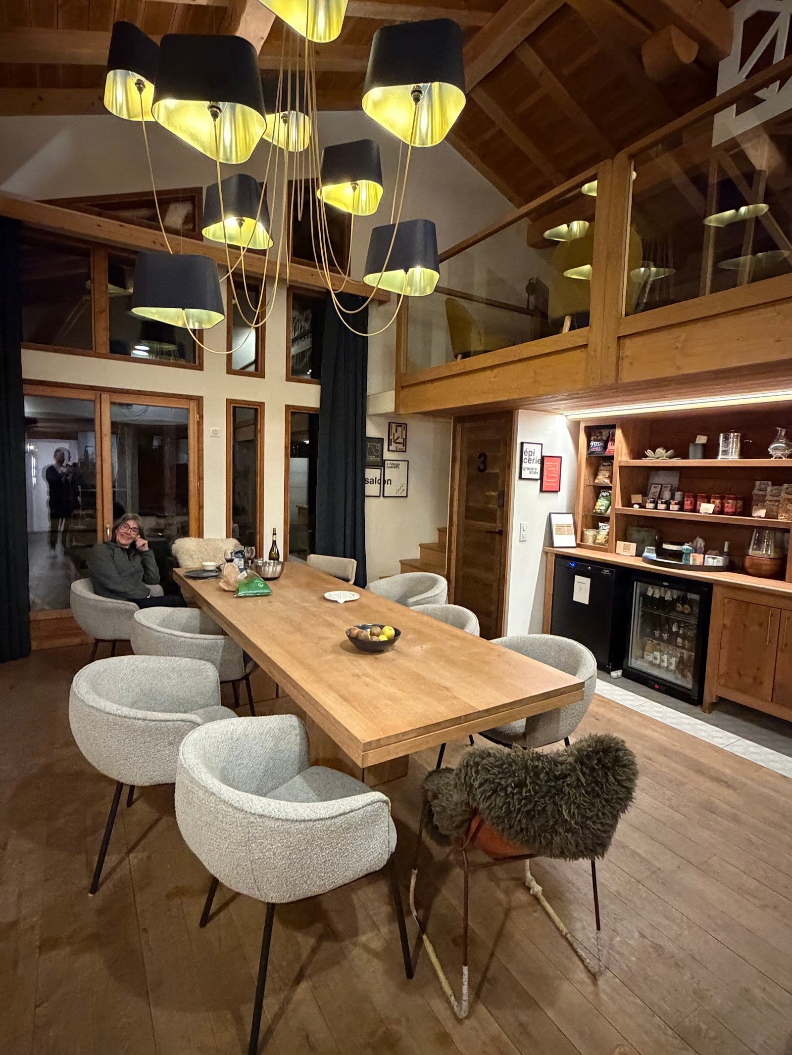 Group of people casually socializing around a large wooden dining table inside a warm, inviting room with high ceilings and wood accents.