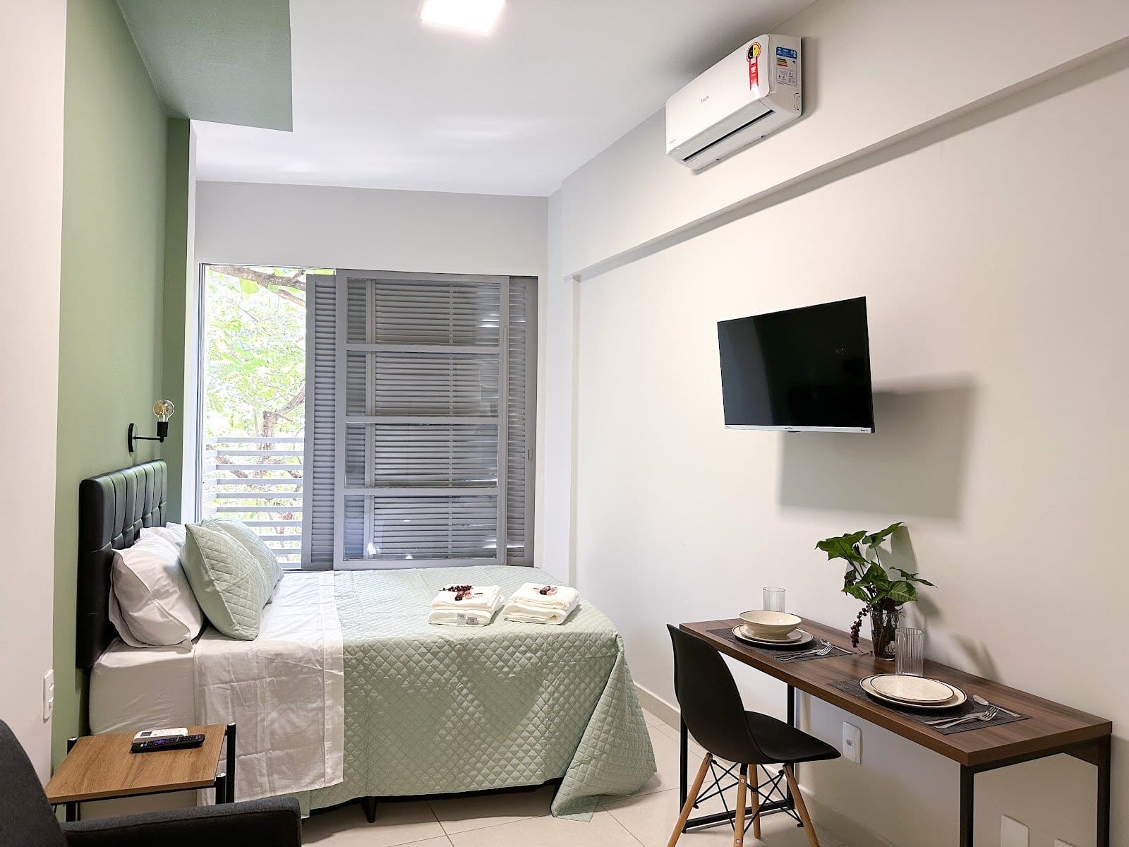 Furnished studio room with green accent wall, double bed and work-and-dining desk at Vivére Coliving in Rio de Janeiro