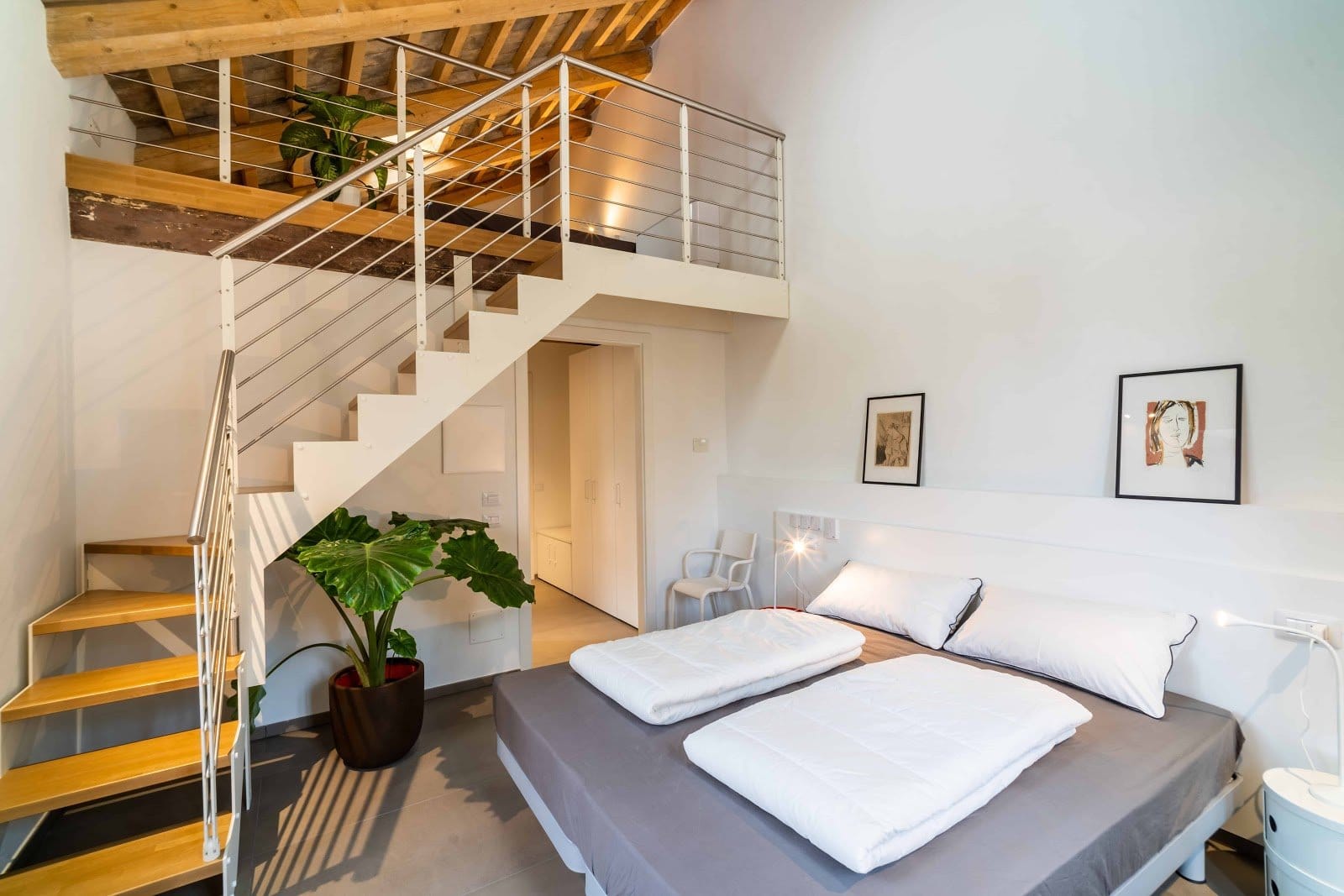 Bright bedroom with large bed, pillows, folded blankets, small staircase to loft, white walls, and framed artwork.