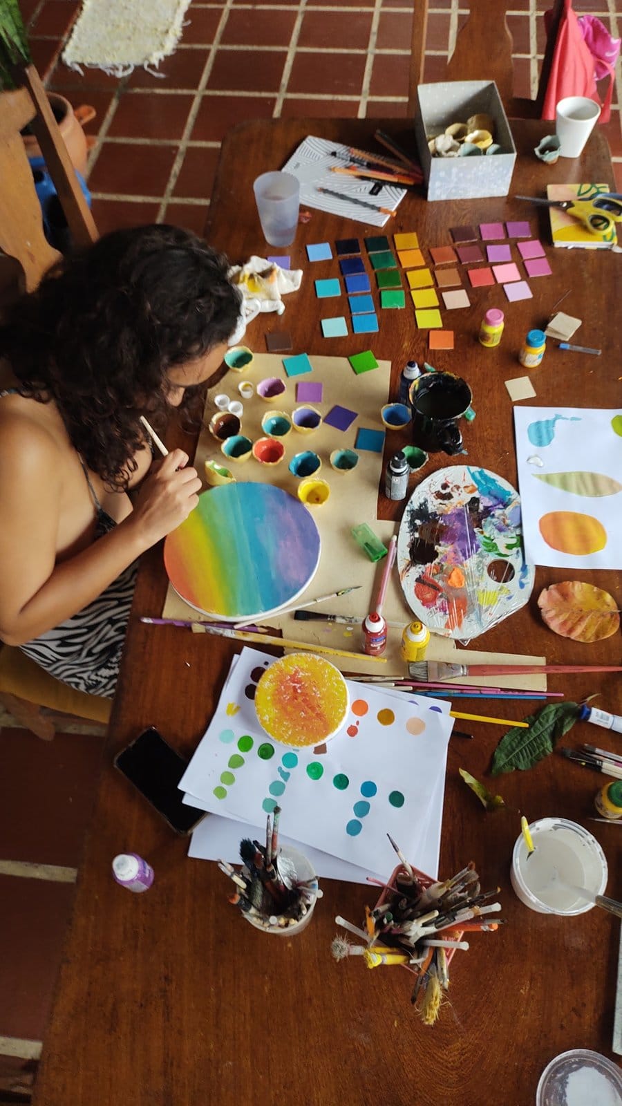 A guest painting a colorful mandala surrounded by art supplies in the ateliê at Tô Aqui Casa de Vivências, Ubatuba