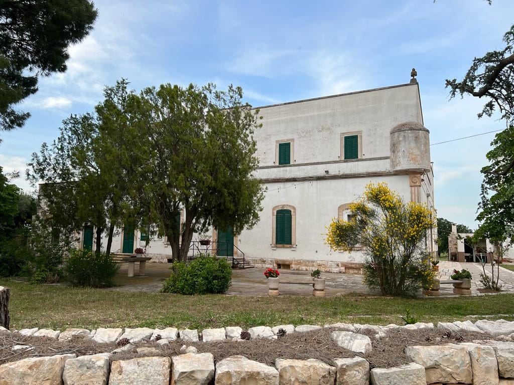 White building with green shutters surrounded by greenery and stone walls under partly cloudy sky.