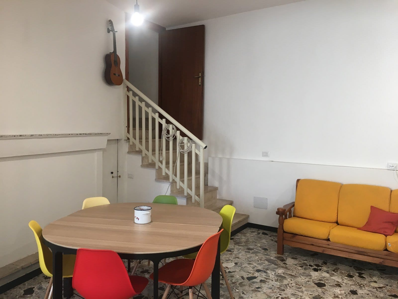 Homely common area with round wooden dining table, colorful red yellow green chairs, orange sofa and guitar on wall at Treballu Rural Coliving in Laconi