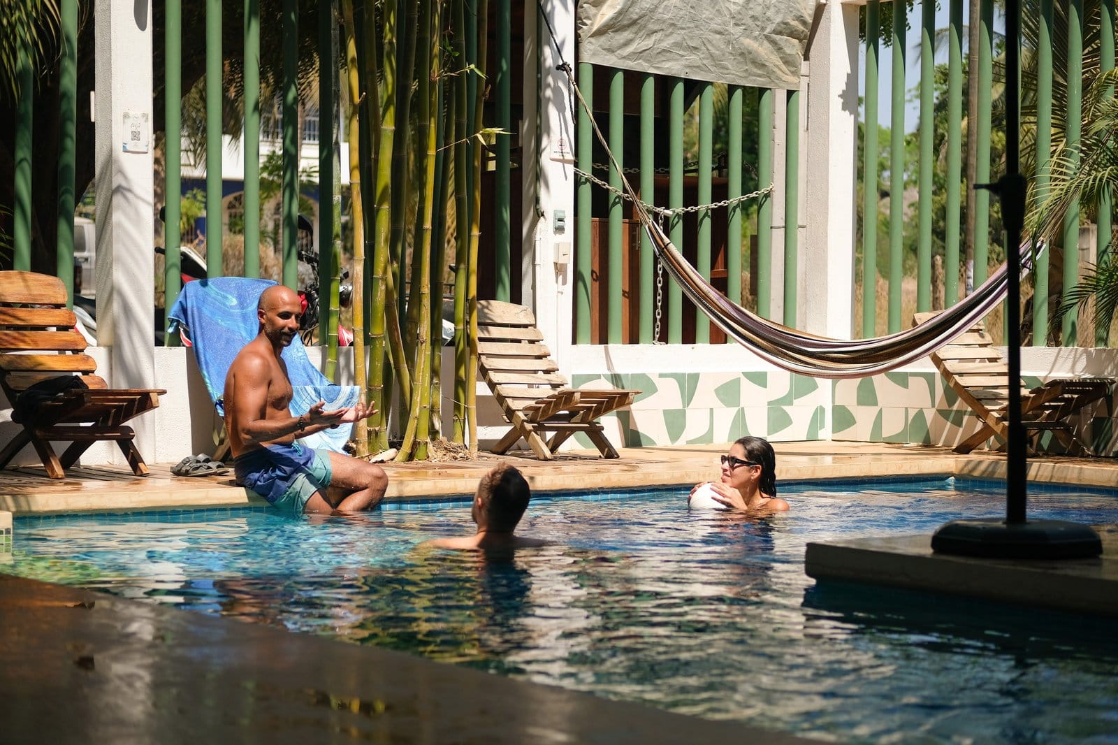 Three Surfbreak PXM residents relaxing by the pool — two in the water, one sitting poolside reading, with hammock and bamboo behind