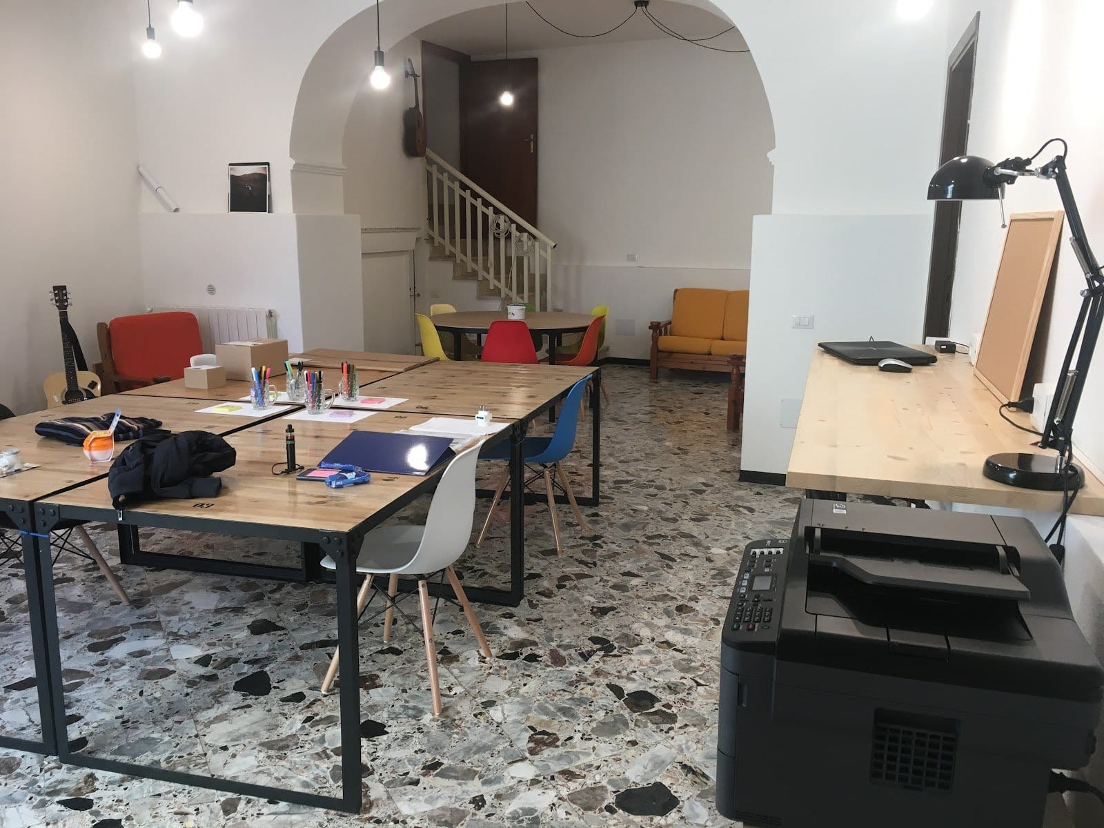 Inviting creative workspace and lounge at Treballu Rural Coliving featuring long wooden desks with colorful chairs, guitars, yellow sofa, arched doorways and historic terrazzo floors in Laconi, Sardinia
