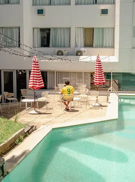 Man in yellow shirt sitting alone next to pool with tables and umbrellas in background.