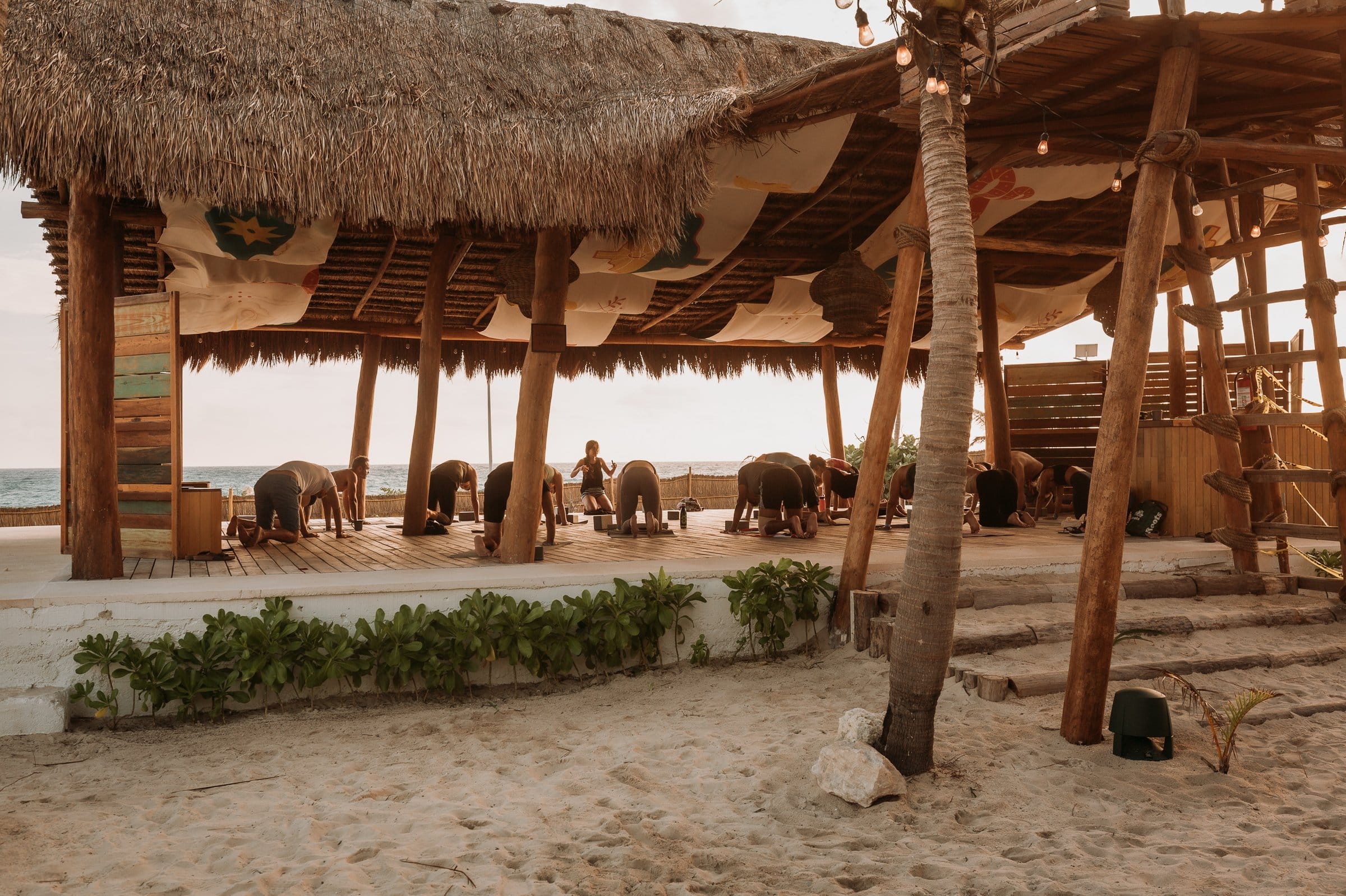 Group yoga class under the open-air thatched beach palapa at Selina Isla Mujeres with the Caribbean sea visible in the background