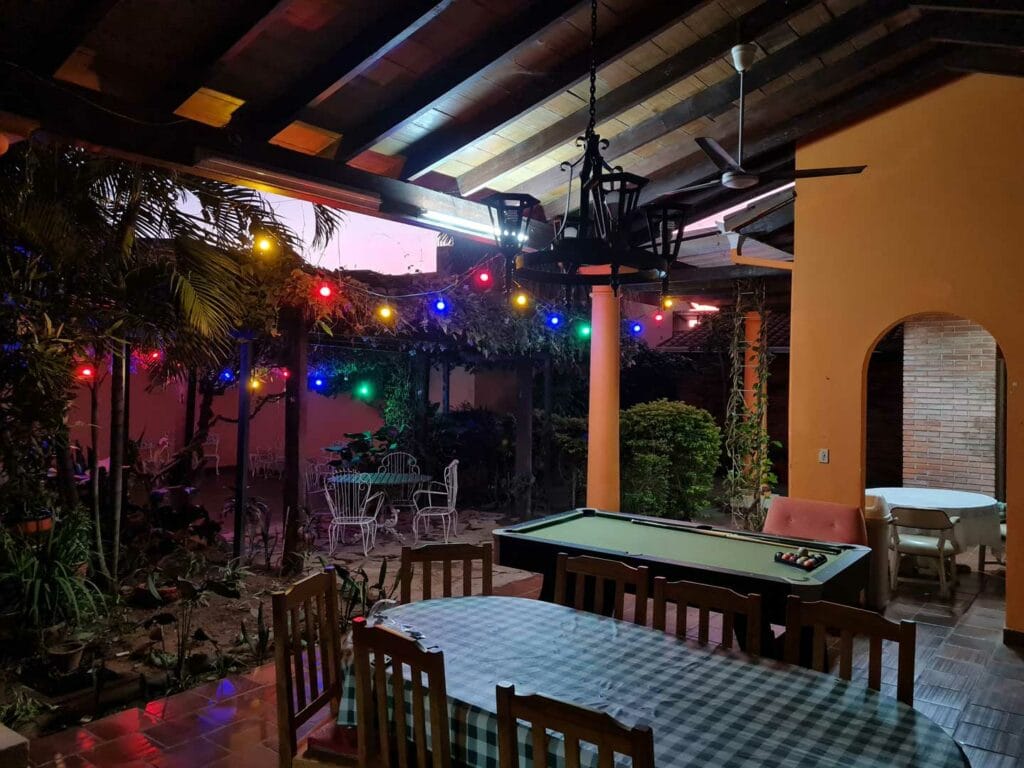 Outdoor covered patio with wooden ceiling beams, string lights, a pool table, and a dining table with chairs surrounded by brick walls and plants.