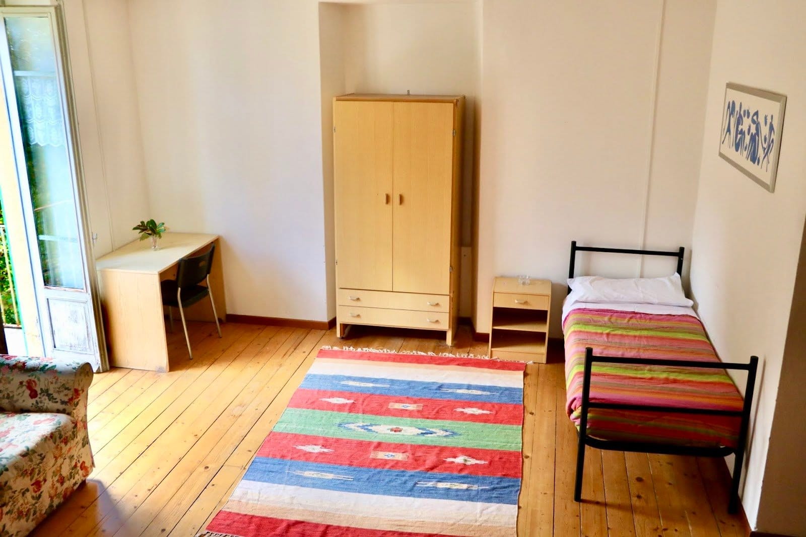 Simple bedroom with single bed, wooden floor, side furniture, and white walls.