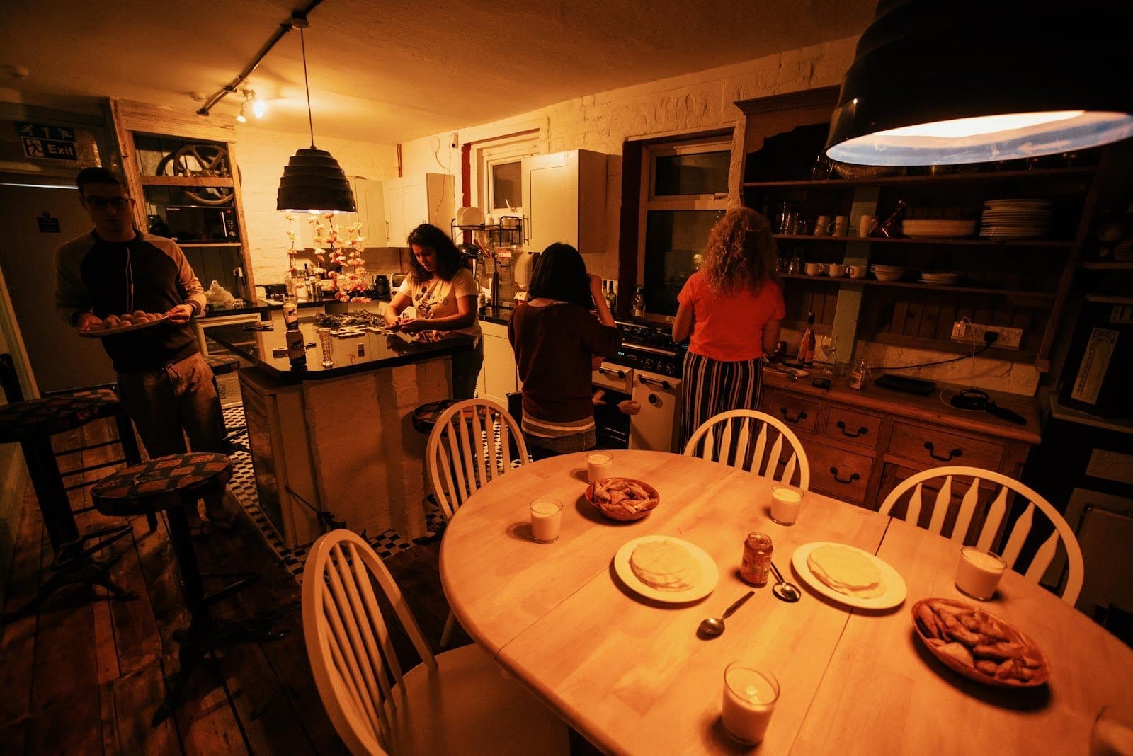 Warmly lit kitchen with four people preparing food and interacting around a wooden table