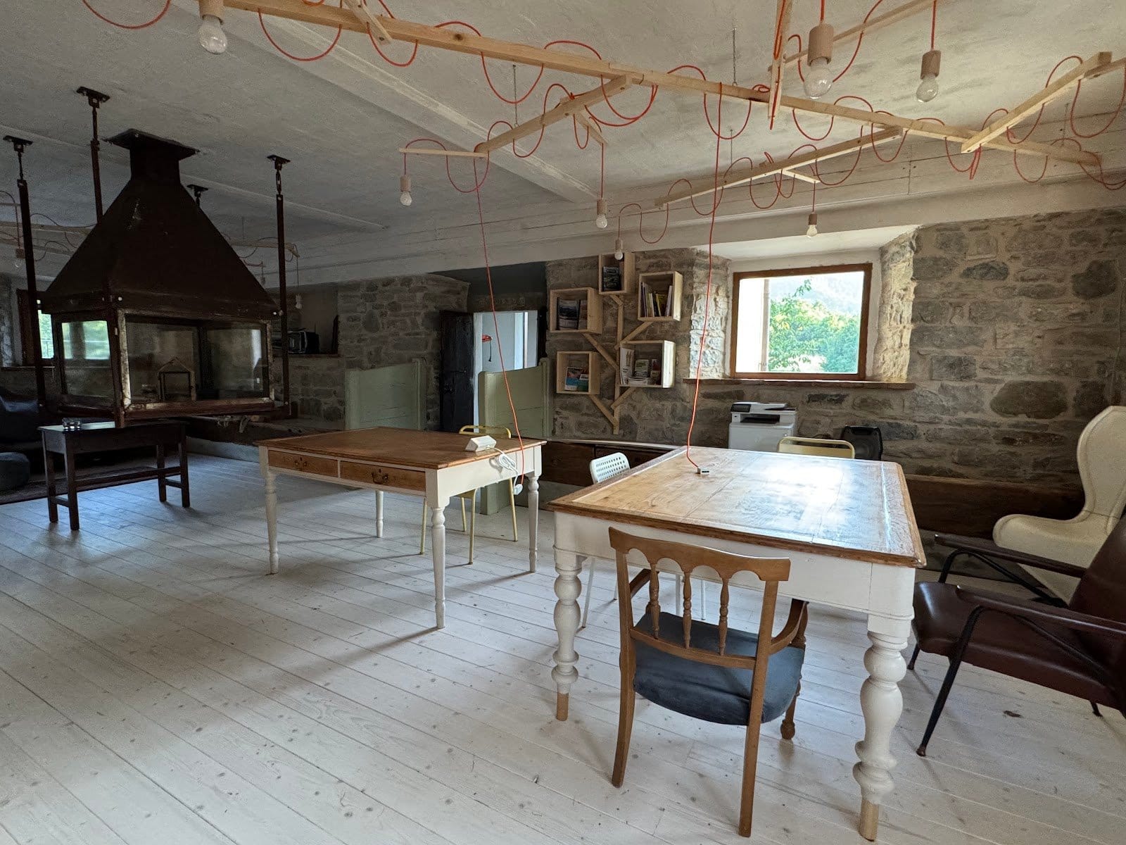 Indoor communal space with wooden floor, two tables, chairs, bookshelves, and a large fireplace, no people visible.