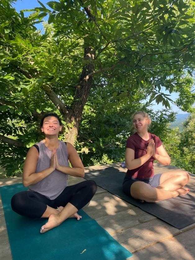 Two people sitting cross-legged on yoga mats in a shaded outdoor setting practicing yoga or meditation.