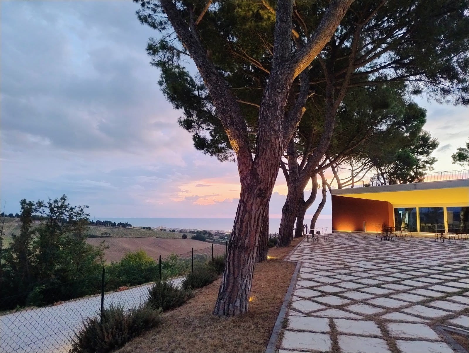 Outdoor terrace area with stone tile flooring, tables and chairs, and large trees with hillside and ocean view at sunset.