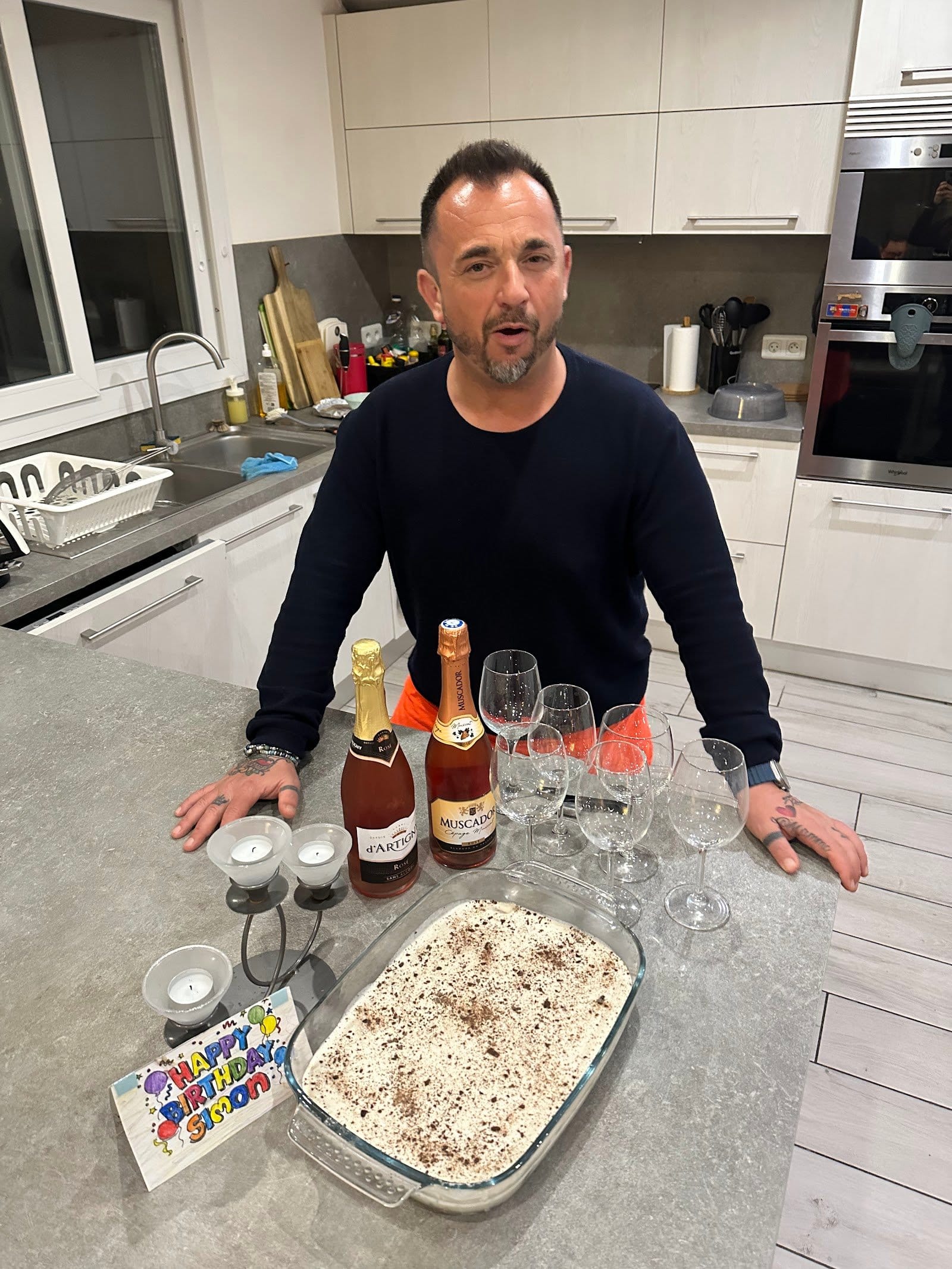 Man stands behind kitchen counter with wine bottles, glasses, and a dessert in a modern kitchen.