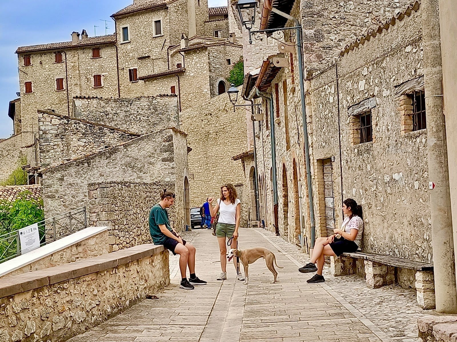 Three people and a dog hanging out on a cobblestone path beside stone buildings