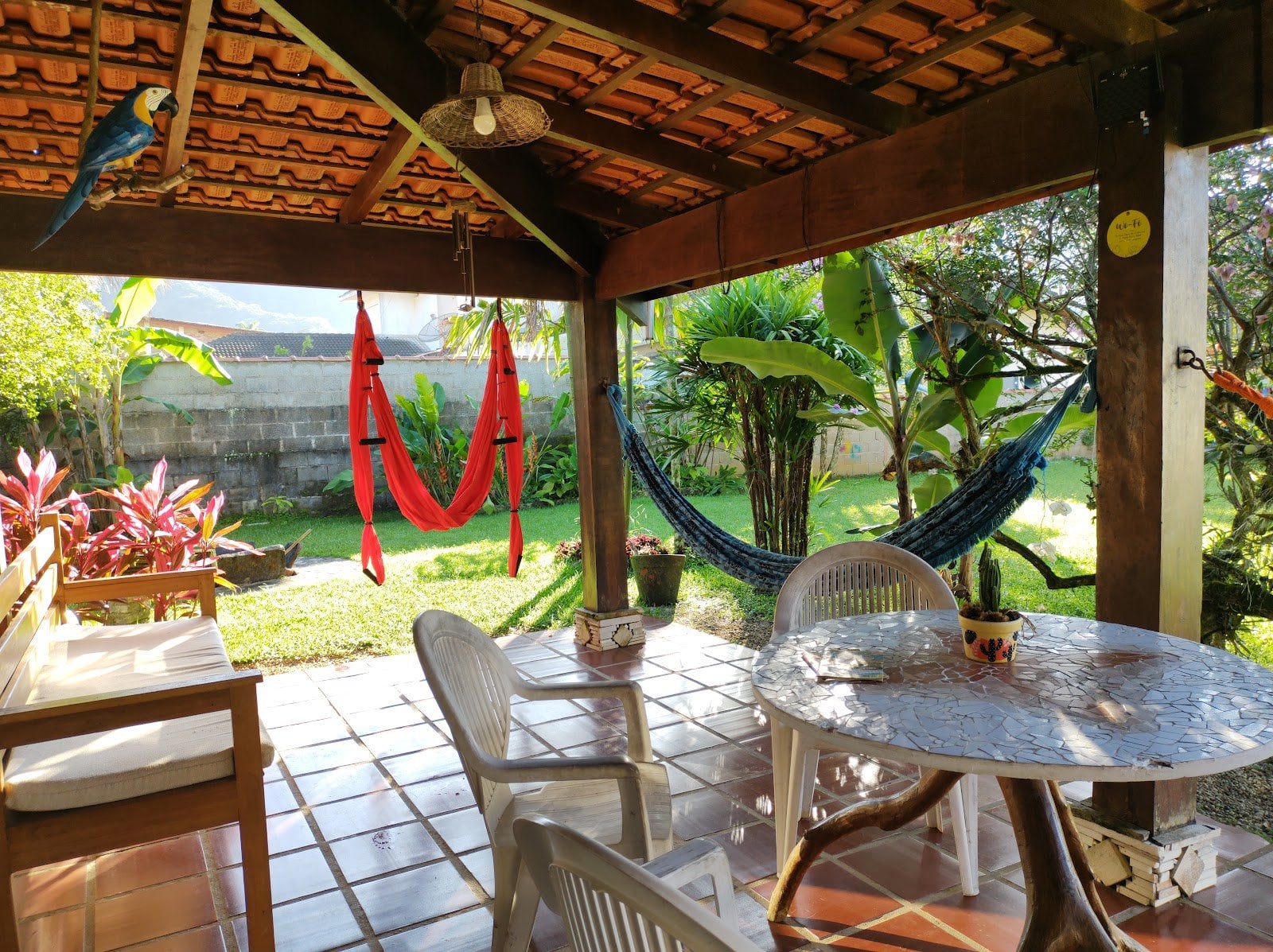 Covered outdoor patio with hammocks, dining tables and a resident parrot overlooking the tropical garden at Tô Aqui Casa de Vivências, Ubatuba
