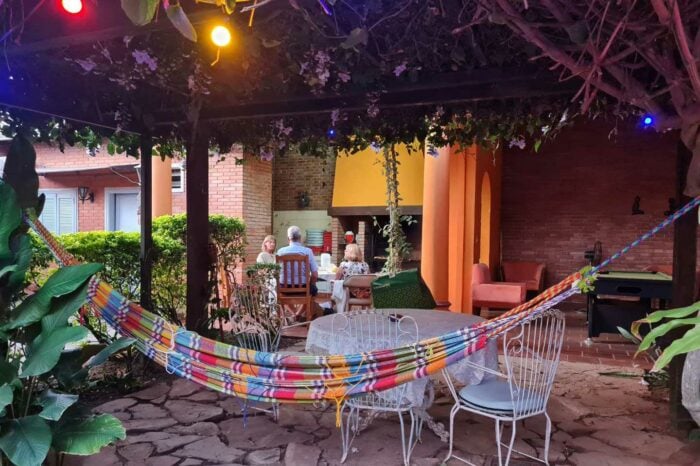 Outdoor seating area with a colorful hammock and a group of people sitting at a table under leafy canopy with string lights.