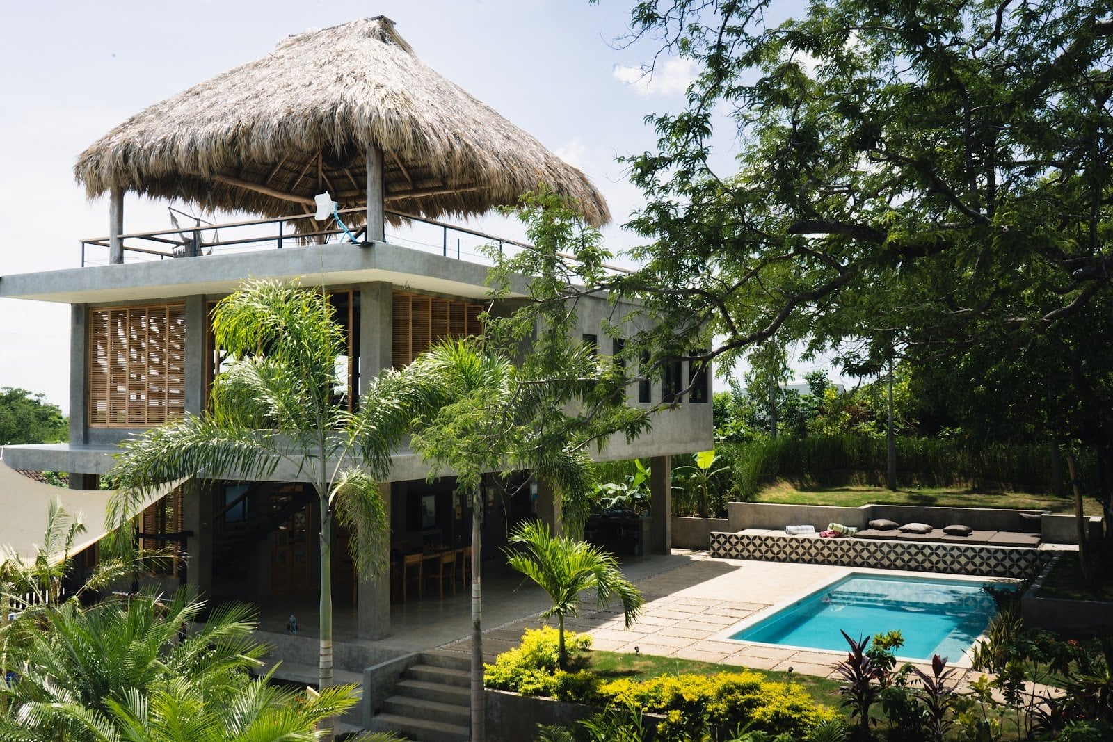 Daylight exterior showing modern two-story building with balconies, pool, trees and garden areas visible