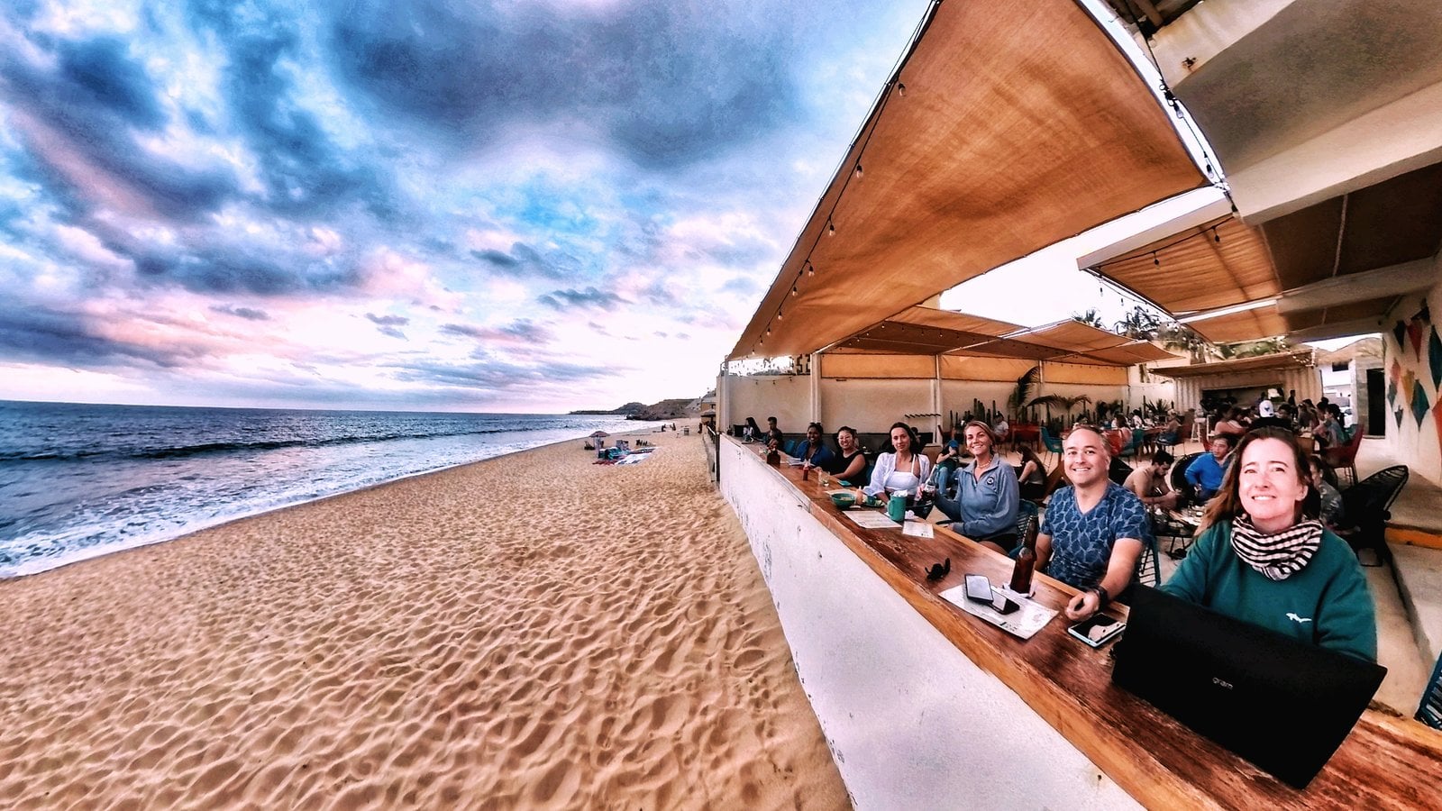 Outsite Los Cabos guests working and dining at a beachside workspace at sunset — panoramic Pacific coast view