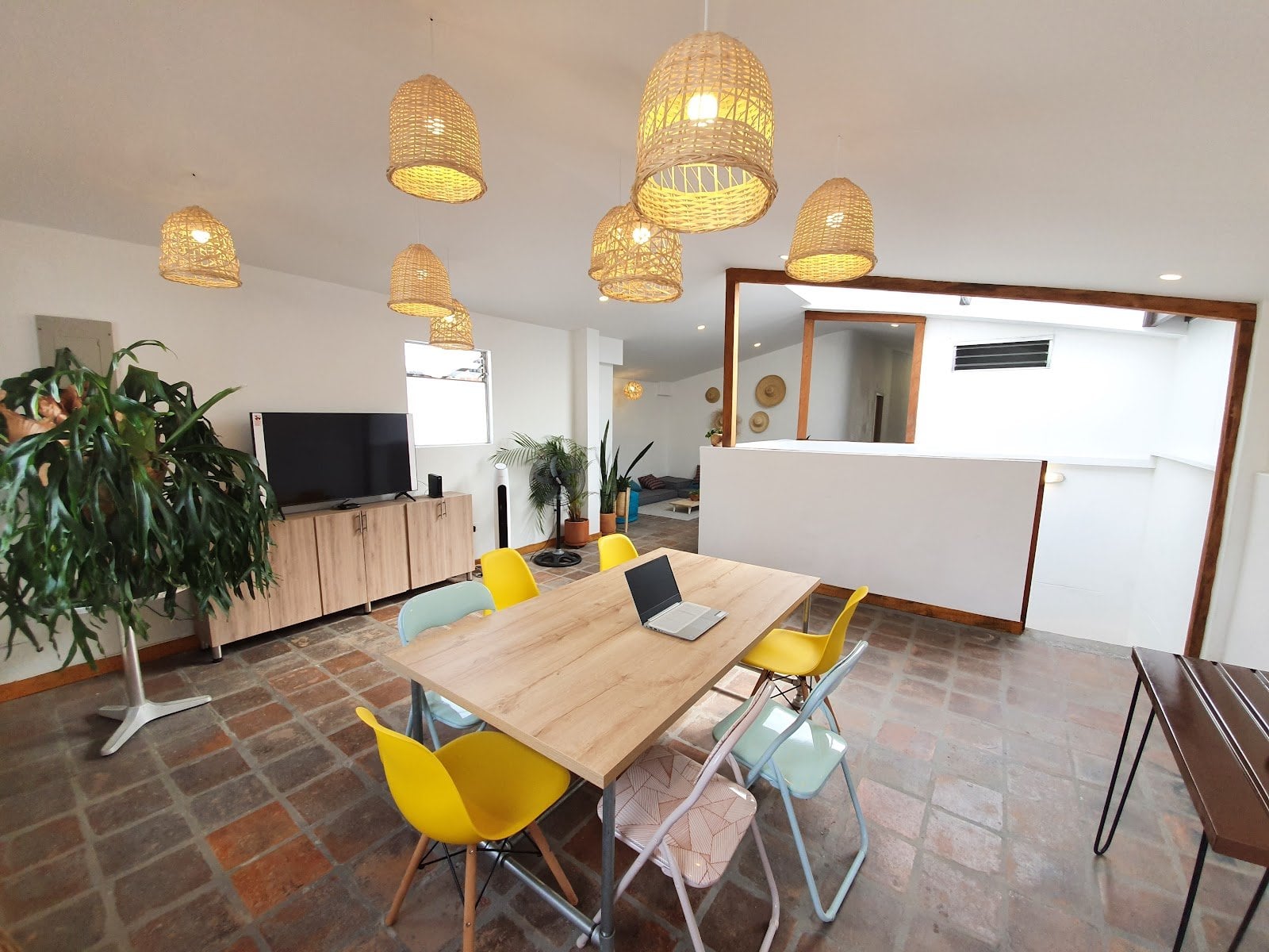 Tribu Coliving sunlit communal dining and lounge area with woven pendant lights, plants and terracotta tile floor in Envigado