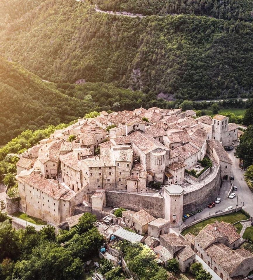 Aerial view of a historic village or town surrounded by hills and greenery under sunlight.