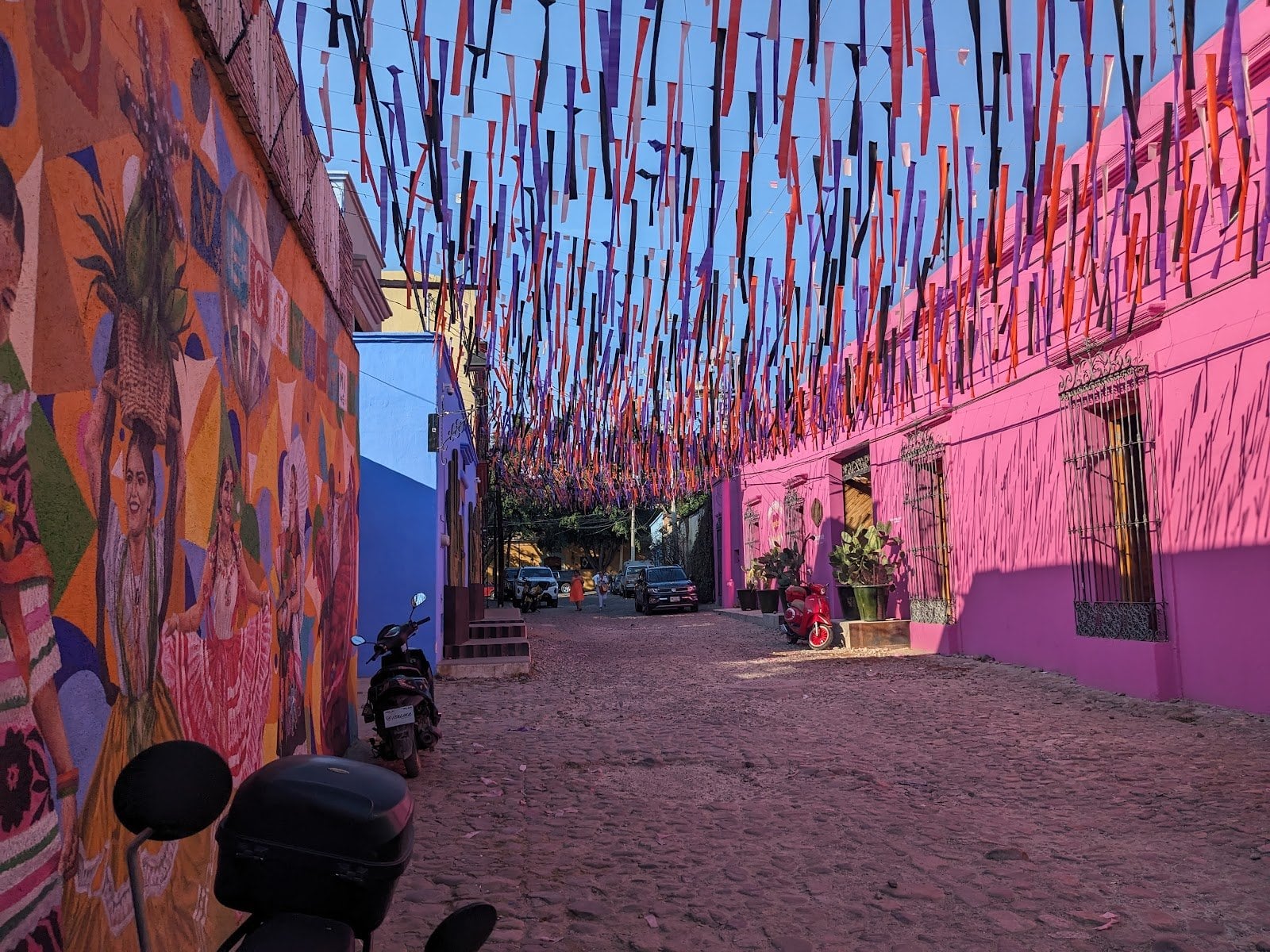 Cobblestone alleyway outside Outsite Oaxaca City at golden hour — the property's pink facade on the right, vibrant mural on the left, and decorative ribbons strung overhead