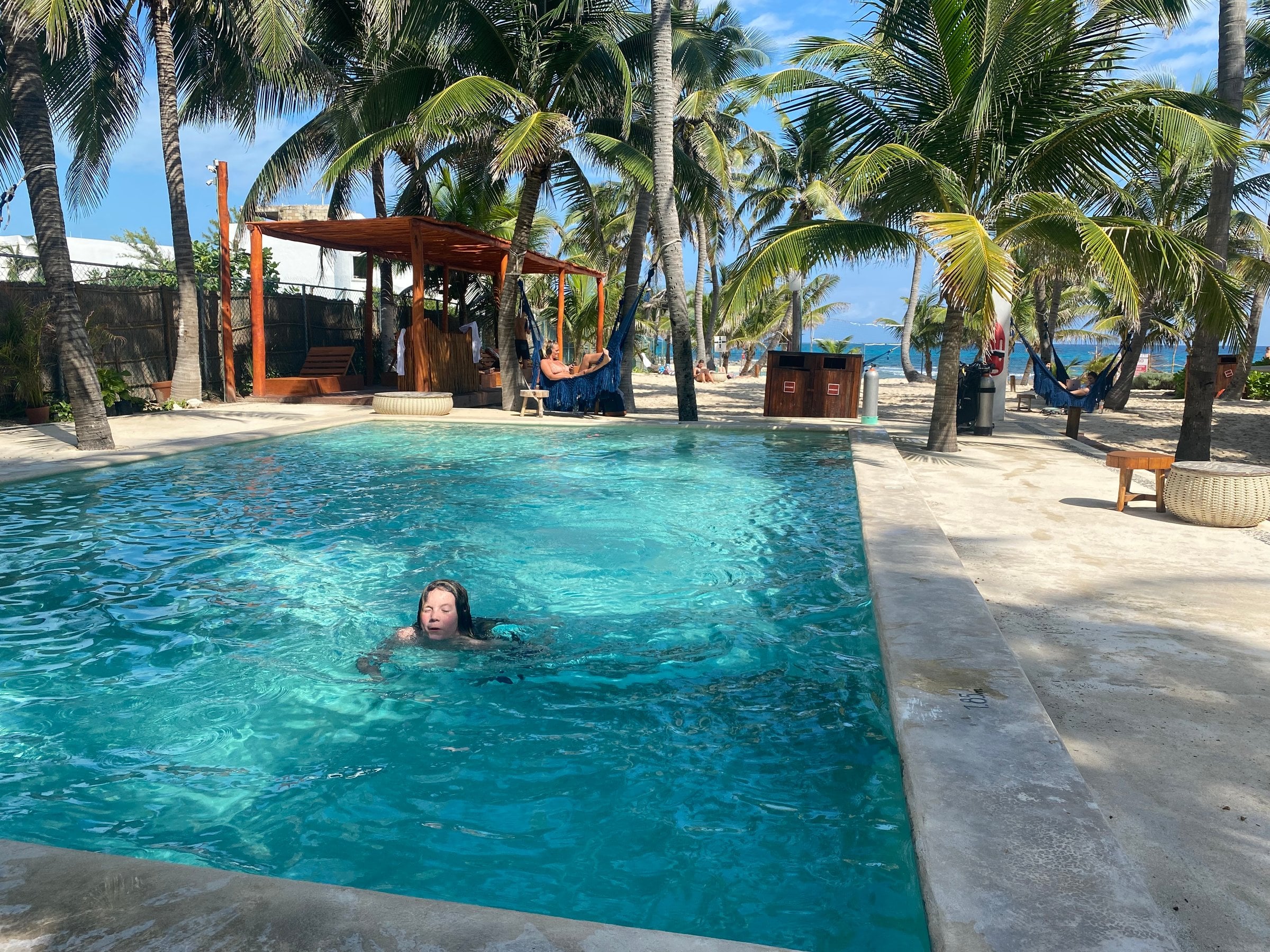 Guest swimming in the turquoise pool at Selina Isla Mujeres with hammocks, palm trees and the Caribbean sea in the background