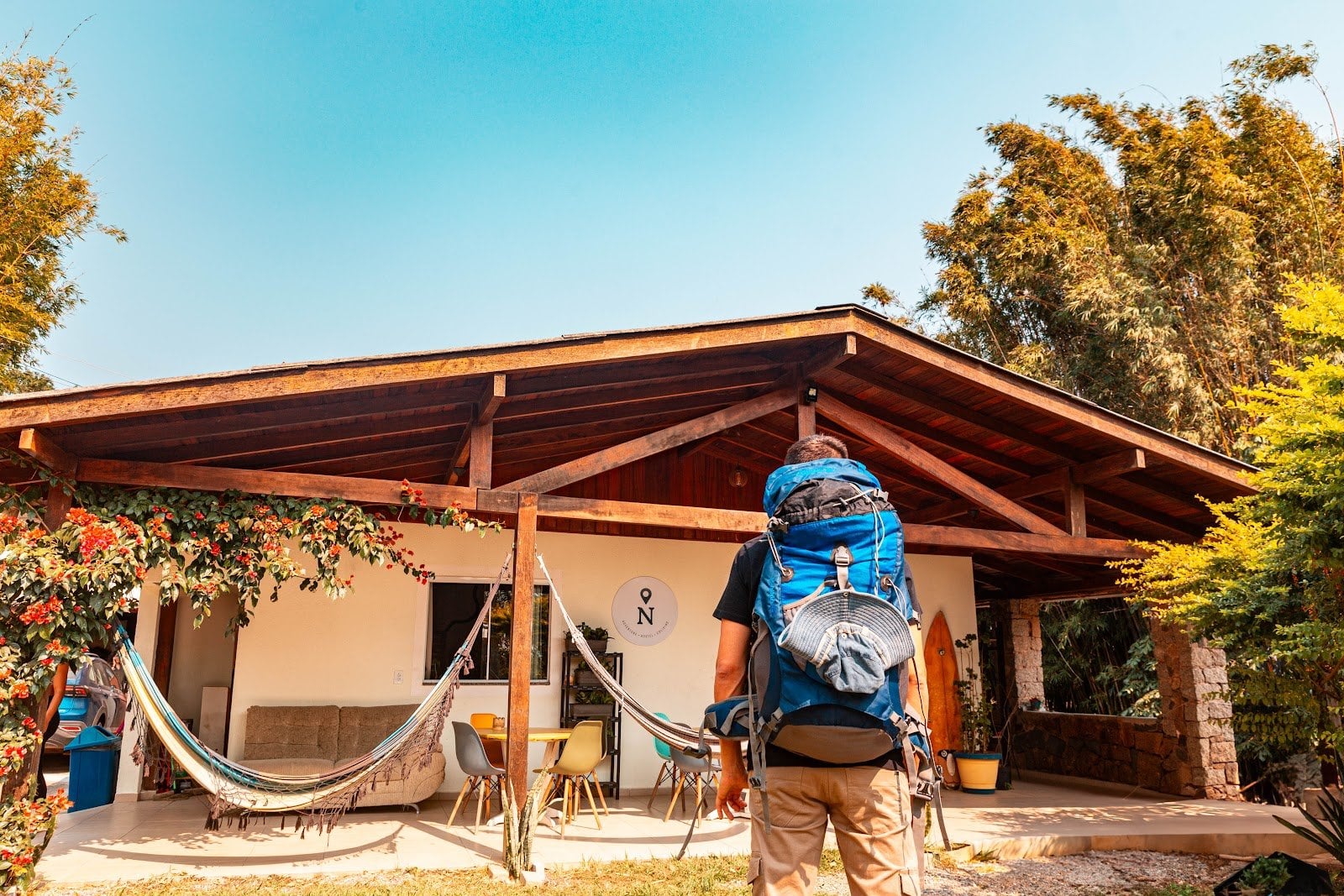 A traveler with a backpack arrives at the hammock-strung front porch of Nômades Riverside hostel in Florianópolis, framed by lush trees in golden light