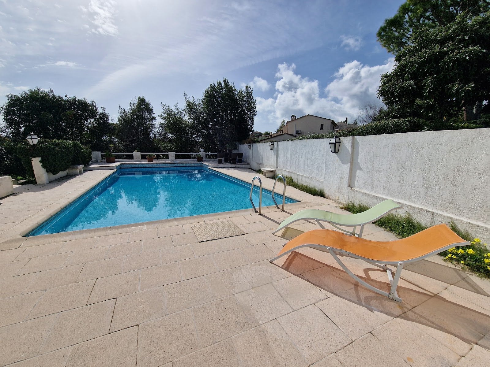 Clear pool with two orange sun loungers on tiled deck surrounded by trees and plants.