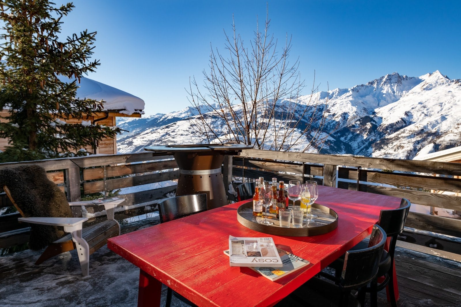 A red outdoor table with drinks and magazines on a snowy terrace overlooking mountain peaks under a clear blue sky with wooden fencing and seating around.