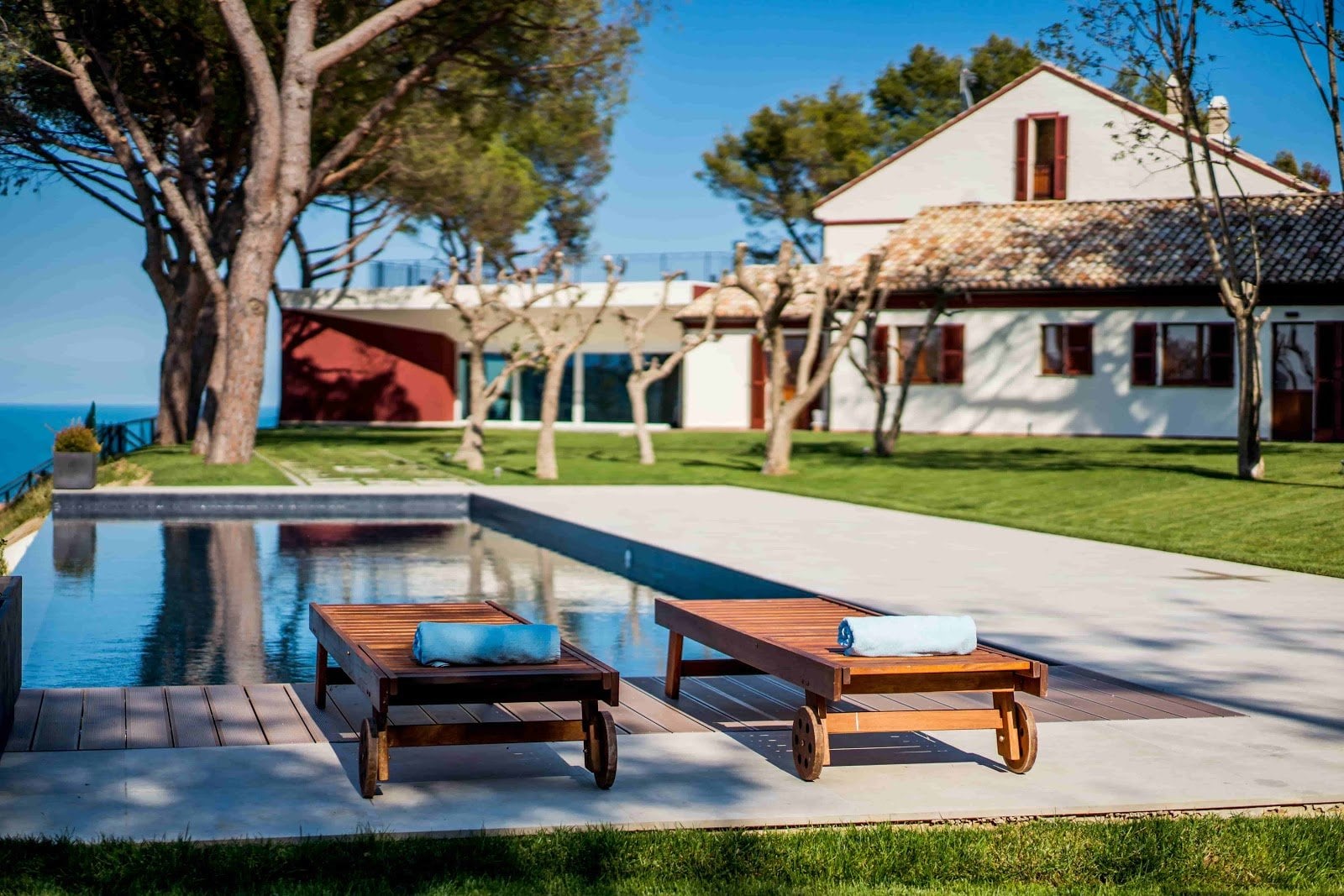 Outdoor pool area with two wooden loungers with rolled towels, grass and trees, white building in background under blue sky.